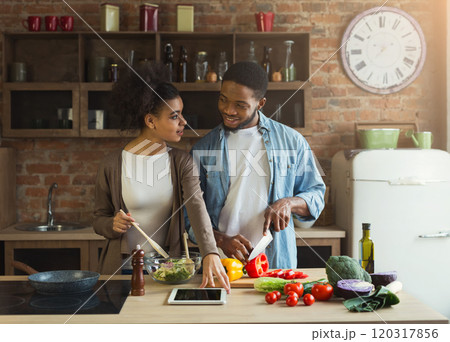 Happy african-american couple cooking healthy food and having fun together in their loft kitchen at home. Woman and man drinking wine. Preparing vegetable salad. Happy african-american couple cooking healthy food and having fun together in their loft kitchen at home. Woman and man drinking wine. Preparing vegetable salad. 120317856