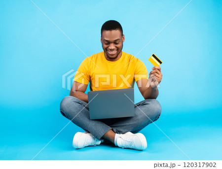 Online Shopping. African American Man Using Laptop Holding Credit Card Purchasing Things In Internet Shop Sitting Over Blue Studio Background. E-Commerce Technologies Concept Online Shopping. African American Man Using Laptop Holding Credit Card Purchasing Things In Internet Shop Sitting Over Blue Studio Background. E-Commerce Technologies Concept 120317902