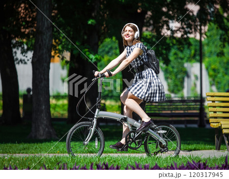 Teen student girl or young woman in plaid dress rides a folding bicycle and listens music in white earphones in the park 120317945