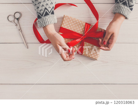 DIY hobby. Woman's hands wrapping christmas or other holiday handmade present in paper with red ribbon. Making bow at present box, decoration of gift. Scissors on white wooden table, top view. 120318063