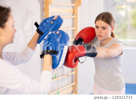 Concentrated teen girl in boxing gloves practicing punches on mitts in hands of instructor 120321715