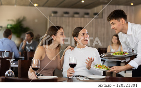 Yyoung waiter serving fresh vegetable salads to female guests visiting restaurant for dinner, sitting at table with glasses of red wine Yyoung waiter serving fresh vegetable salads to female guests visiting restaurant for dinner, sitting at table with glasses of red wine 120321801