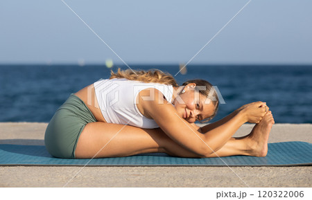 On windy day young slender woman does yoga near sea doing Pashchimottanasana 120322006