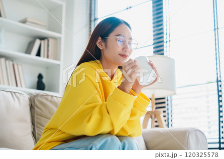 Young asian woman wearing eyeglass sitting on sofa and holding mug in living room at home Young asian woman wearing eyeglass sitting on sofa and holding mug in living room at home 120325578