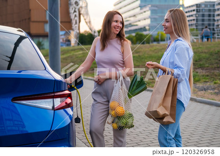 Two women engage in conversation and holding bags with grocery while one charges an electric car in a parking lot 120326958