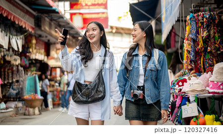 Two happy Asian female tourists explore a local market in Thailand, walking along the street. 120327416
