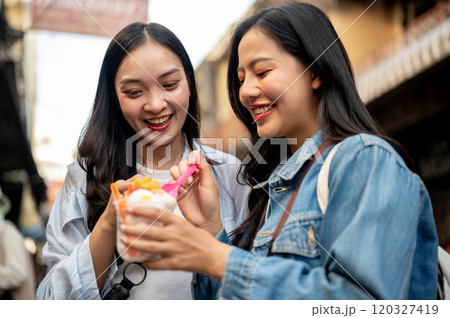 Two pretty, joyful Asian female tourists enjoy street food at a market in Thailand, eating ice cream Two pretty, joyful Asian female tourists enjoy street food at a market in Thailand, eating ice cream 120327419