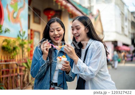 Two pretty, joyful Asian female tourists enjoy street food at a market in Thailand, eating ice cream Two pretty, joyful Asian female tourists enjoy street food at a market in Thailand, eating ice cream 120327422