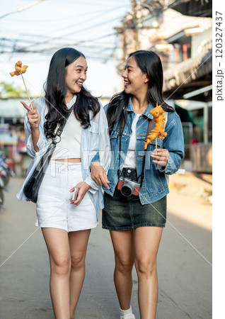 Two happy Asian female friends enjoying a city stroll, eating street food in Thailand on a sunny day 120327437