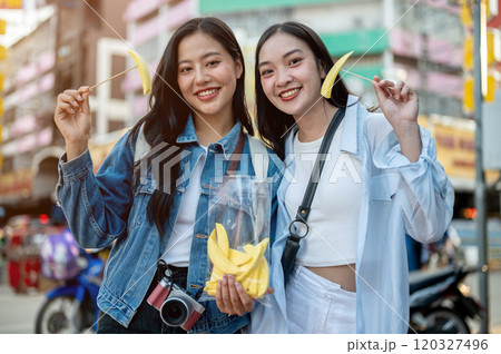 Two happy Asian female tourists enjoy eating while exploring a street food market. Two happy Asian female tourists enjoy eating while exploring a street food market. 120327496