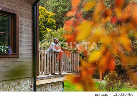Man is enjoying a relaxing, peaceful moment at home, standing on patio, leaning against railing and looking at autumn nature around him. 120330469