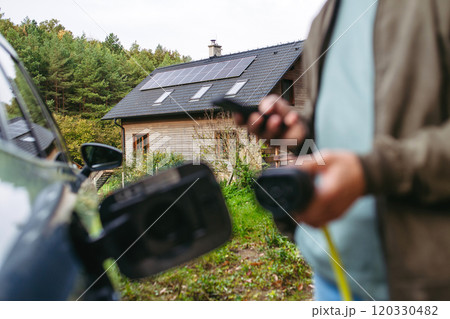 Man charging electric car in front of his house, plugging the charger into the charging port. House with solar panel system on roof behind him. 120330482