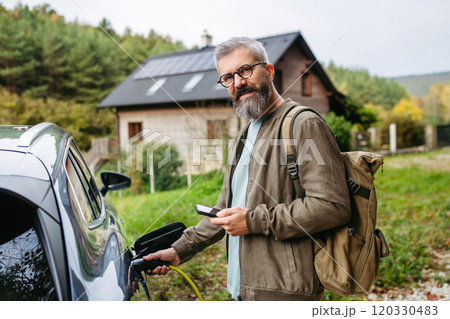 Man charging electric car in front of his house, plugging the charger into the charging port. House with solar panel system on roof behind him. Man charging electric car in front of his house, plugging the charger into the charging port. House with solar panel system on roof behind him. 120330483