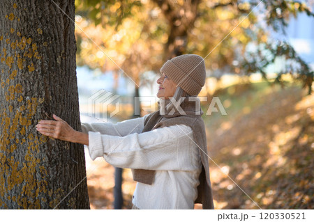 Portrait of a beautiful senior woman hugging tree trunk with closed eyes. Elderly woman in connection with nature. Portrait of a beautiful senior woman hugging tree trunk with closed eyes. Elderly woman in connection with nature. 120330521