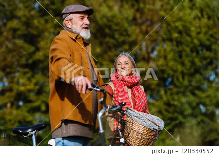 Lovely elderly couple on a walk in the forest, pushing their bikes side by side. Seniors in love on stroll through autumn nature, enjoying a peaceful moment together. 120330552