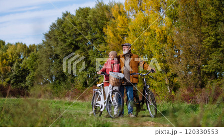 Lovely elderly couple on a walk in the forest, pushing their bikes side by side. Seniors in love on stroll through autumn nature, enjoying a peaceful moment together. 120330553