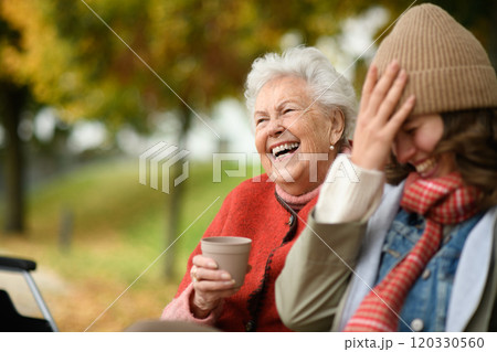 Portrait of a granddaughter and grandmother sitting on bench in autumn park, drinking coffee and laughing together. Portrait of a granddaughter and grandmother sitting on bench in autumn park, drinking coffee and laughing together. 120330560