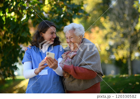 Portrait of nurse and elderly woman on walk in the park during warm autumn day. Young caregiver looking through photos of senior patient's family on a phone. 120330582