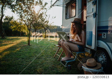 Young beautiful woman is on a camping trip in nature, sitting in front of caravan with notebook on knees. Caravan as homeoffice, working remotely. 120330613