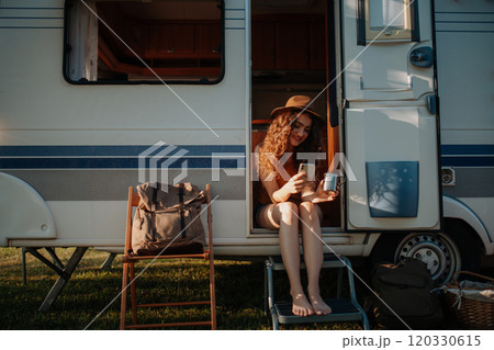 Portrait of young beautiful woman on a camping trip in nature, sitting on caravan steps with smartphone and coffee in hand. 120330615