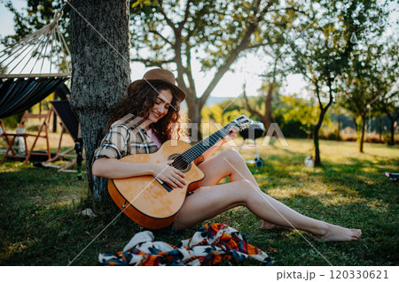 Young beautiful woman playing guitar outside, sitting on the ground and leaning against tree. Solo camping trip in nature. Young beautiful woman playing guitar outside, sitting on the ground and leaning against tree. Solo camping trip in nature. 120330621