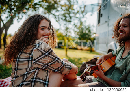 Couple sitting by caravan, enjoying peaceful moment. Man playing guitar and singing song to his girlfriend. Camping trip for young people. Couple sitting by caravan, enjoying peaceful moment. Man playing guitar and singing song to his girlfriend. Camping trip for young people. 120330622