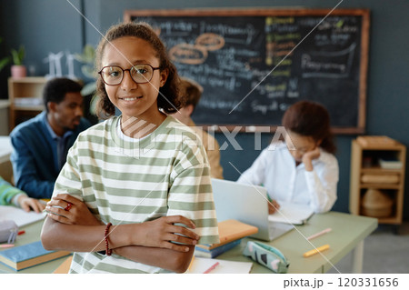 Waist up portrait of teenage African American boy wearing glasses while standing with arms crossed in school classroom and smiling at camera, copy space Waist up portrait of teenage African American boy wearing glasses while standing with arms crossed in school classroom and smiling at camera, copy space 120331656