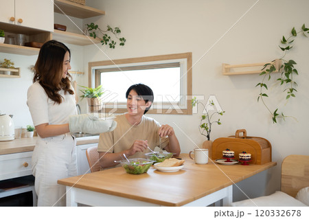 Young woman wearing an oven mitt serving a freshly prepared dish to her husband in a minimalist kitchen Young woman wearing an oven mitt serving a freshly prepared dish to her husband in a minimalist kitchen 120332678