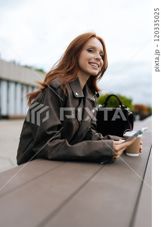 Vertical portrait of cheerfully woman with flowing red hair drinking coffee and checking phone while standing outside by river. Happy redhead female smiling in calm outdoor setting 120335025