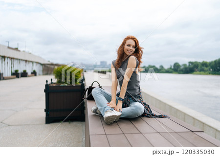 Portrait of happy female finding serenity on river pier, savoring tranquil atmosphere and cloudy sky. Pretty smiling young woman appears at ease, taking respite from urban living outdoors. 120335030