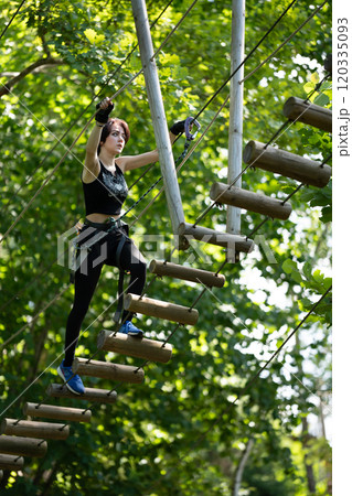 A woman confidently navigates her way across a high ropes course, surrounded by lush, vibrant greenery all around her 120335093