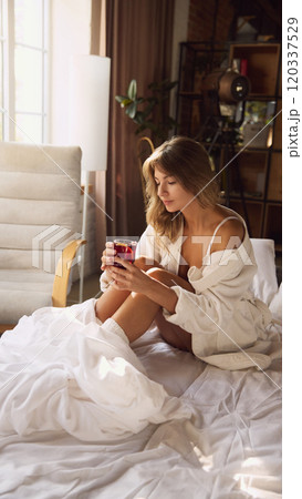 Woman in white bathrobe sits on bed, drinking mulled wine on bedroom. Calm moment of relaxation in natural light. Woman in white bathrobe sits on bed, drinking mulled wine on bedroom. Calm moment of relaxation in natural light. 120337529
