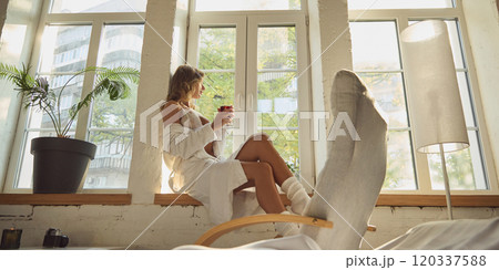 Woman in white lingerie and robe sits on windowsill with glass of red drink, enjoying relaxed moment with sunlight filtering through window. 120337588