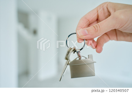 Close up hand holding keys with house shaped keychain in front of blurred new apartment interior. Concept of mortgage and new house purchase. Real estate transactions 120340507