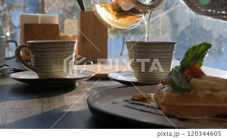 Close-up of a young woman pouring steaming herbal tea, savoring a peaceful breakfast with family in a winter forest setting 120344605