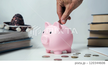 A hand places a coin into a pink piggy bank on a white table surrounded by scattered coins and a crumpled dollar bill, illustrating savings, financial security, and budgeting 120344606