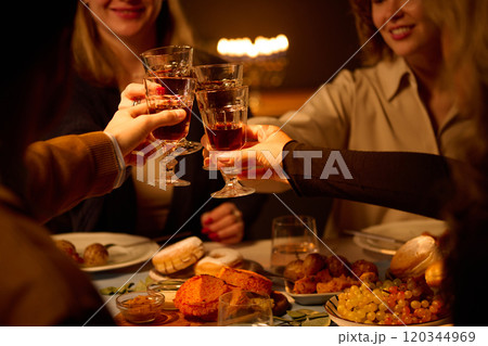 Medium close up of unrecognizable friends clinking glasses with red wine while sitting at table in dimly lit room 120344969