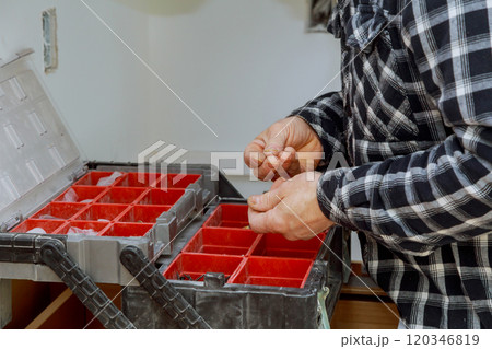 Man sorts through small components in toolbox with red containers, focusing on organizing materials at workshop setting. Man sorts through small components in toolbox with red containers, focusing on organizing materials at workshop setting. 120346819