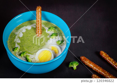 Vibrant green soup is served in blue bowl, garnished with sliced boiled eggs, broccoli florets, alongside pretzel sticks on dark surface. Vibrant green soup is served in blue bowl, garnished with sliced boiled eggs, broccoli florets, alongside pretzel sticks on dark surface. 120346822