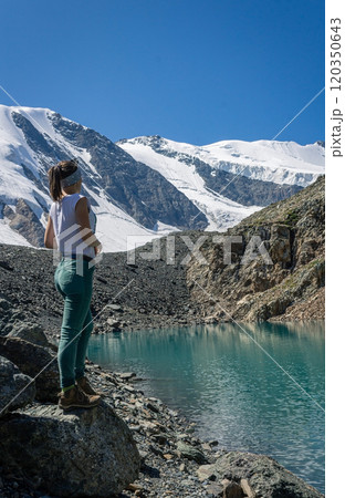 Woman stands on the shore of a turquoise lake, against the backdrop of high mountains with snow-capped peaks Woman stands on the shore of a turquoise lake, against the backdrop of high mountains with snow-capped peaks 120350643