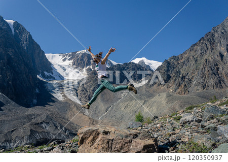 Young woman in a jump against the backdrop of the high mountains of Altai with snow-capped peaks and a glacier. 120350937