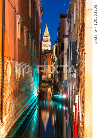 A narrow canal in Venice, Italy, at night, with a boat and a tall bell tower of St Mark's Campanile in the distance 120353238