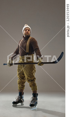 Full-length portrait of man, hockey player in sweater, pants with suspenders and knitted hat, standing with stick in grey studio background 120353449