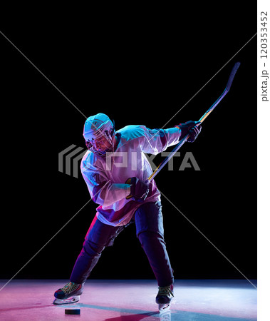 Competitive man, hockey player wearing full game gear, in motion with stick, competing for the victory against black studio background in neon light 120353452