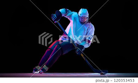 Focused man, professional hockey player in white gear in motion with stick, controlling puck against black studio background in neon light Focused man, professional hockey player in white gear in motion with stick, controlling puck against black studio background in neon light 120353453