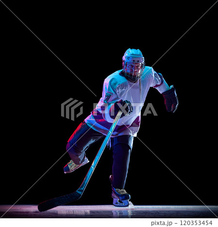 Dynamic image of competitive men's, hockey player in white sportswear in motion on ring with stick, against black studio background in neon light 120353454