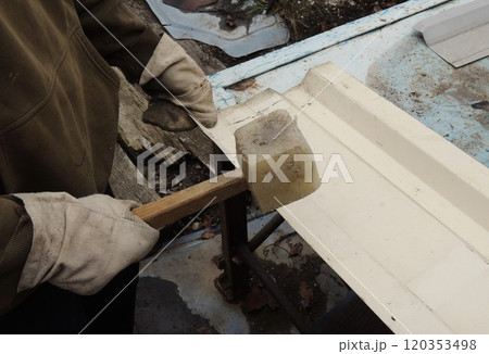 A man on the street, wearing work clothes and gloves, repairs or straightens a sheet of metal with a wooden mallet. A man on the street, wearing work clothes and gloves, repairs or straightens a sheet of metal with a wooden mallet. 120353498