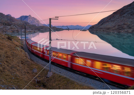 Moving red train near azure mountain lake, snowy rocks at sunset Moving red train near azure mountain lake, snowy rocks at sunset 120356871