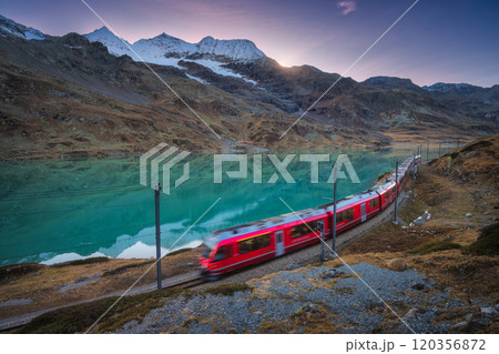 Moving red train near azure mountain lake, snowy rocks at sunset 120356872