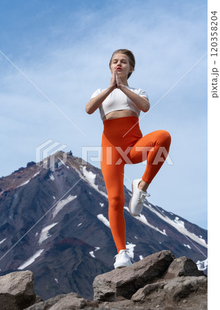 Woman doing yoga with one leg up against background of volcano. She's got great body and is really into Zen meditation with eyes closed. Adult Gen Y yoga instructor in t-shirt, yoga pants and trainers Woman doing yoga with one leg up against background of volcano. She's got great body and is really into Zen meditation with eyes closed. Adult Gen Y yoga instructor in t-shirt, yoga pants and trainers 120358204